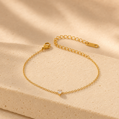 A delicate gold chain bracelet with a small clear heart-shaped gemstone, displayed on a textured beige stone surface with soft shadows.
