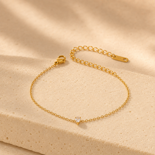 A delicate gold chain bracelet with a small clear heart-shaped gemstone, displayed on a textured beige stone surface with soft shadows.